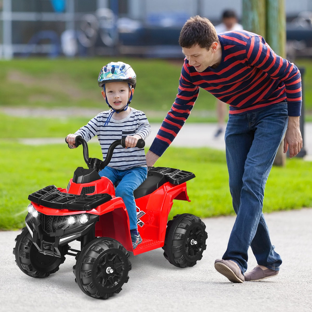 The kids electric car being pushed by a man along a paved path in a park with a child riding