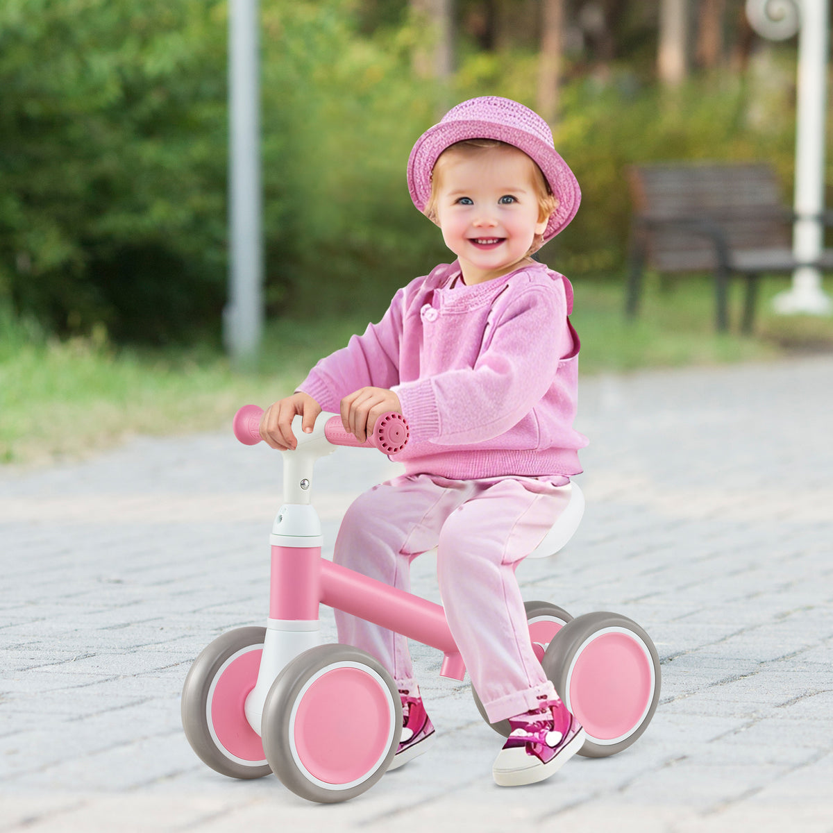 A smiling toddler girl wearing a pink hat and outfit riding the balance bike outdoors