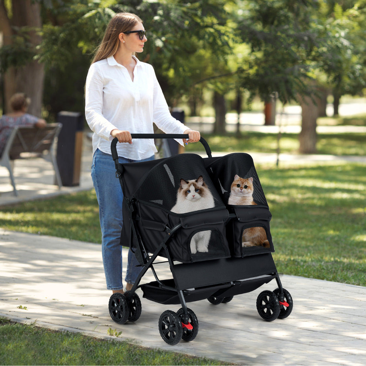A woman pushes a black double dog stroller with two pets inside on a paved path in a park