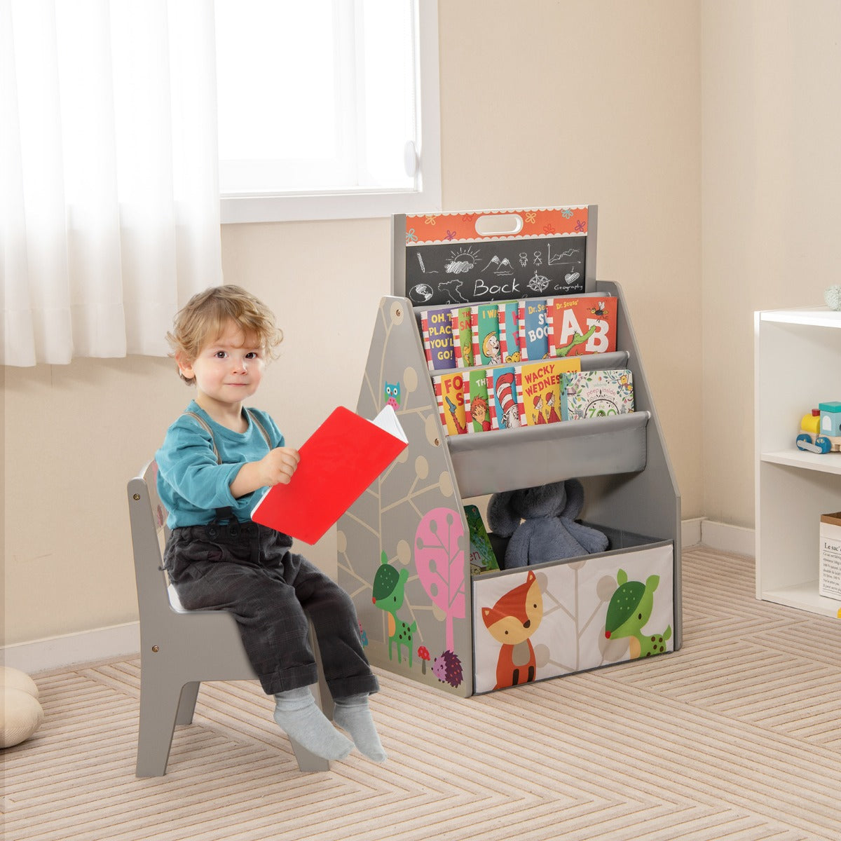 A young boy sits on the grey chair reading a book next to the art easel and bookshelf