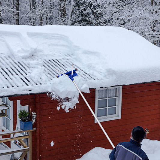 The garden tool being used to clear heavy snow from the roof of a small red wooden cabin