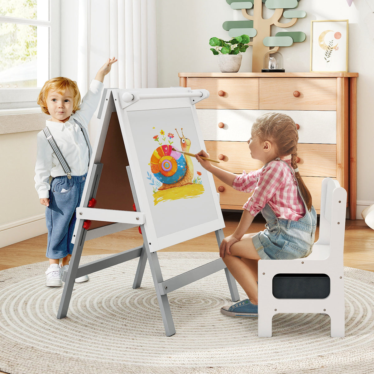 Two children paint and draw on the white easel, one sitting and one standing