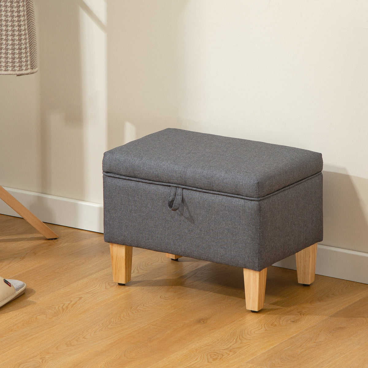 The charcoal grey storage ottoman is placed on a wooden floor next to a coat stand