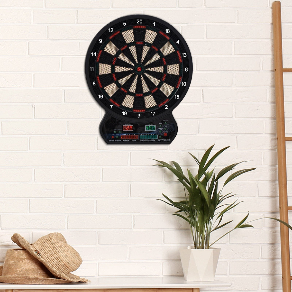 The electronic dartboard mounted on a white brick wall above a table with a plant