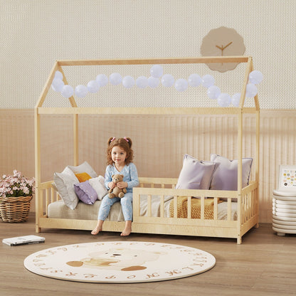 A young girl sits on the natural wood house-shaped kids bed frame in a decorated bedroom