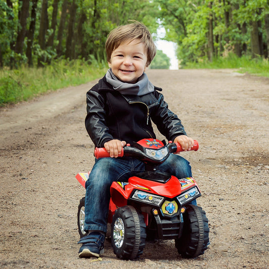 A happy toddler boy wearing a black jacket and grey scarf rides the red quad bike toy on a dirt track