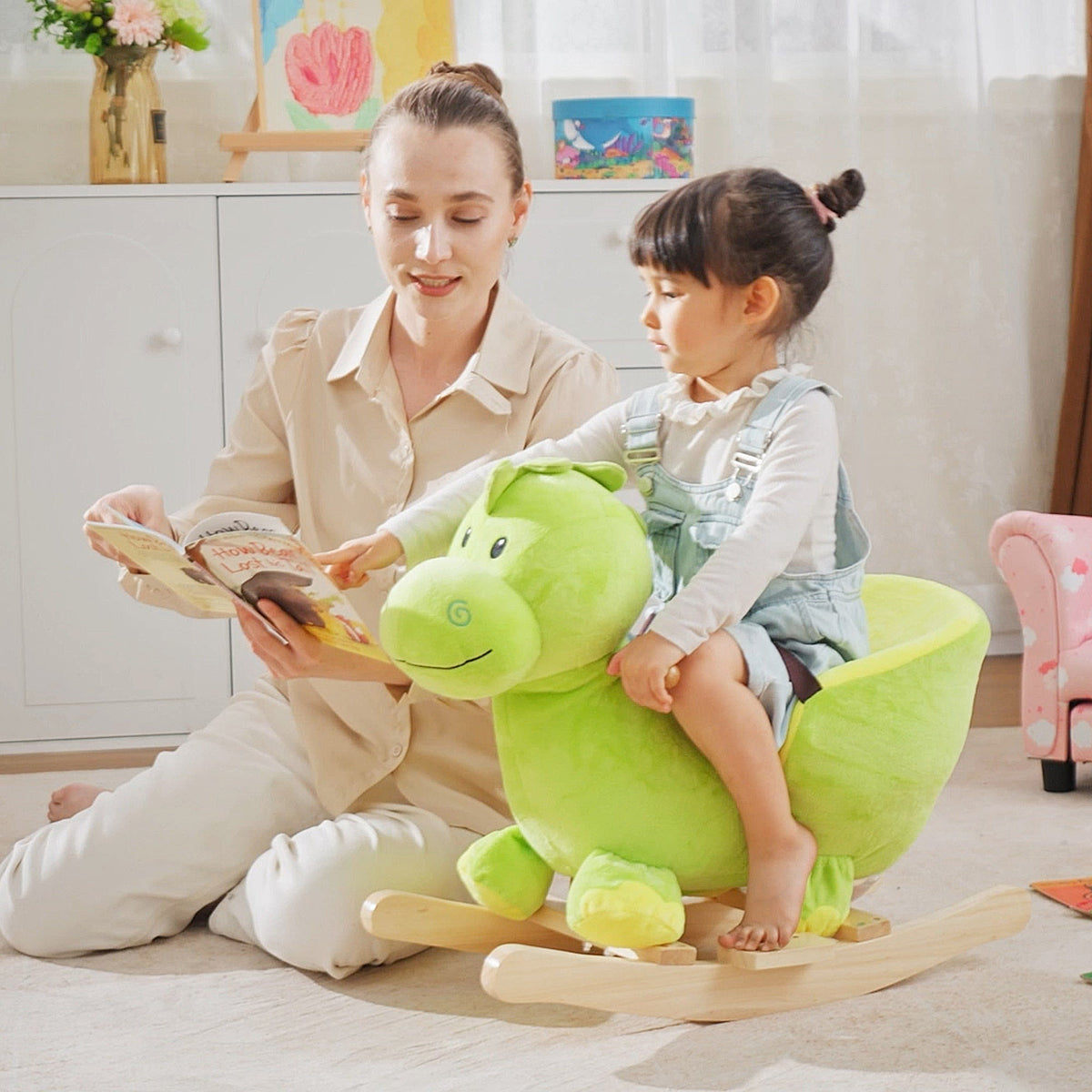 A young child sits on the plush dinosaur rocking horse while a woman reads a book
