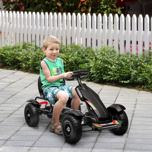 A young boy sitting on the pedal go kart on a paved driveway in front of a white garden fence