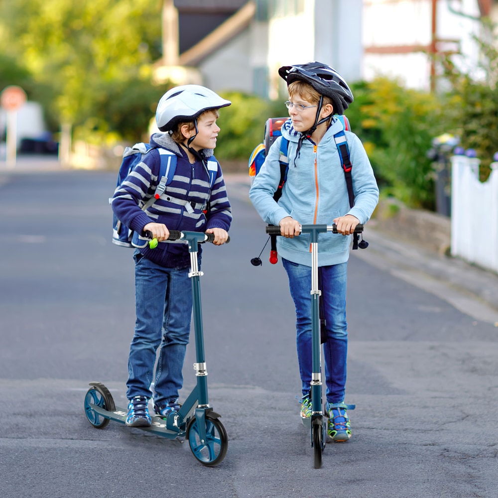 Two boys wearing helmets ride blue kick scooters on a street