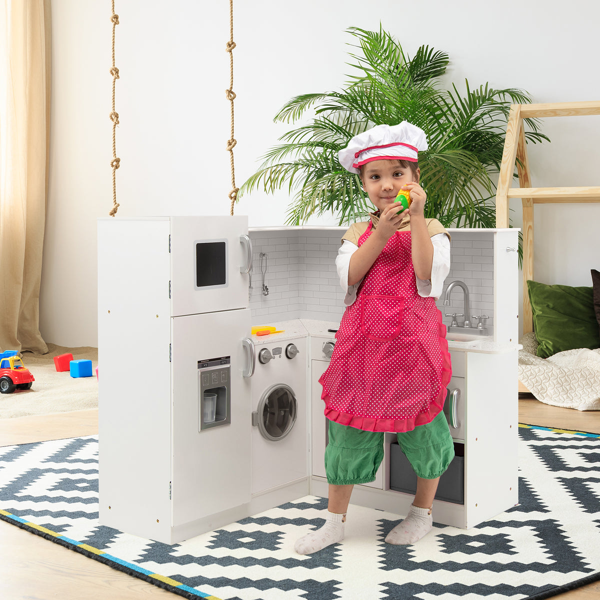 A young child wearing an apron and chef hat plays with a white play kitchen set in a room