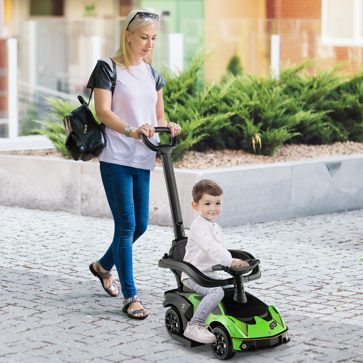 A woman pushes a young boy in the green ride on car along a pavement