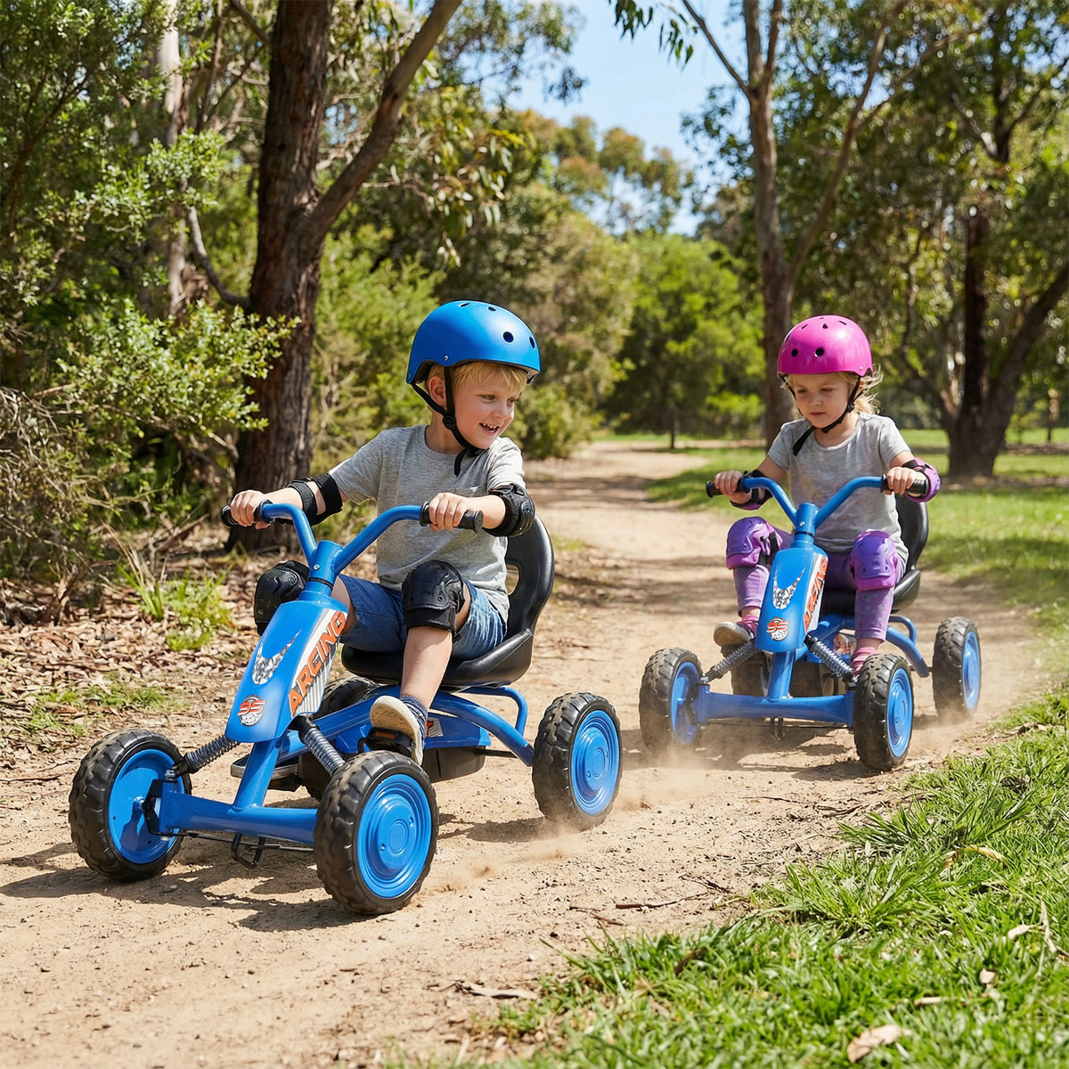 Two children riding the pedal ride on toys along a dirt path in a park with trees