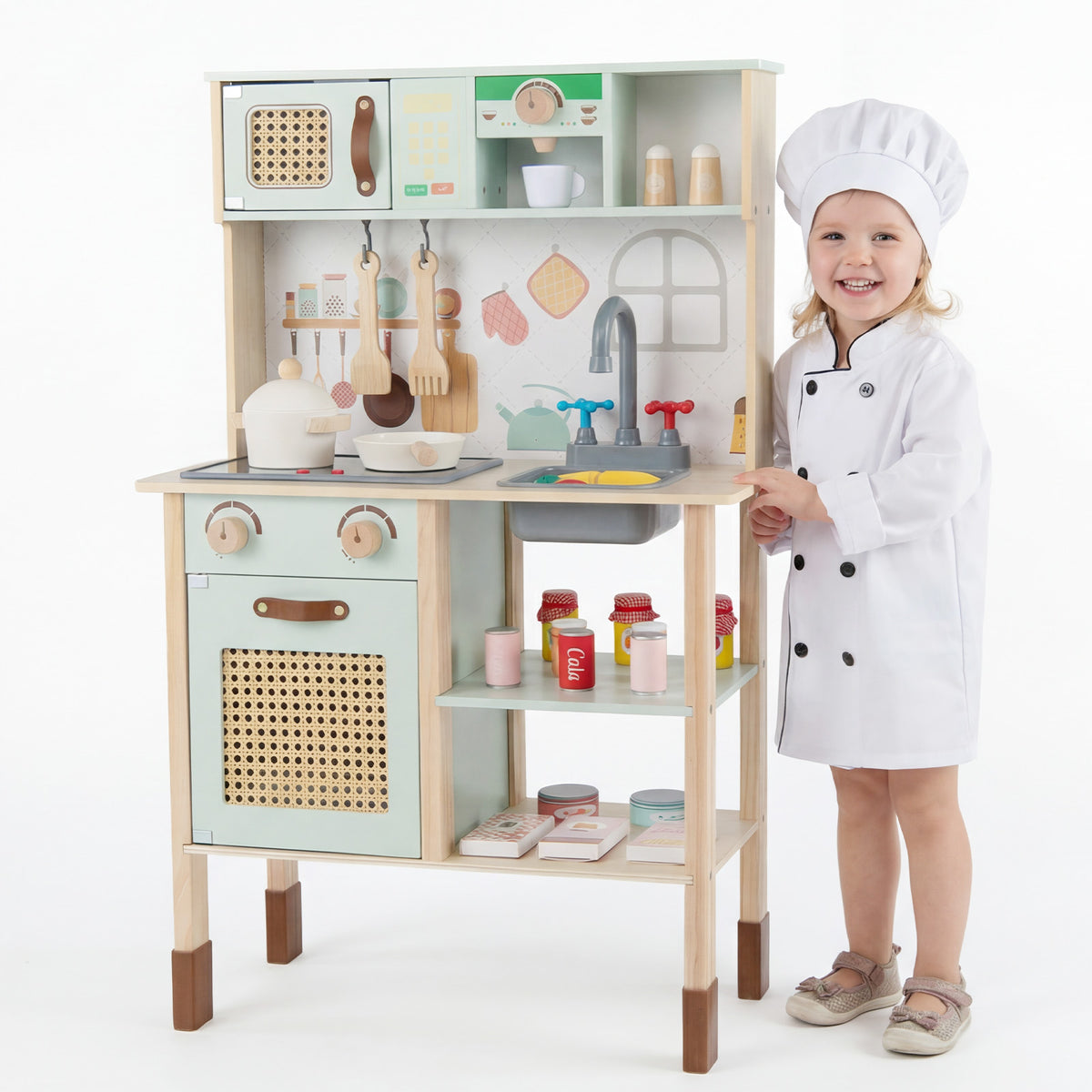 A smiling girl in a chef's outfit stands next to the green wooden play kitchen