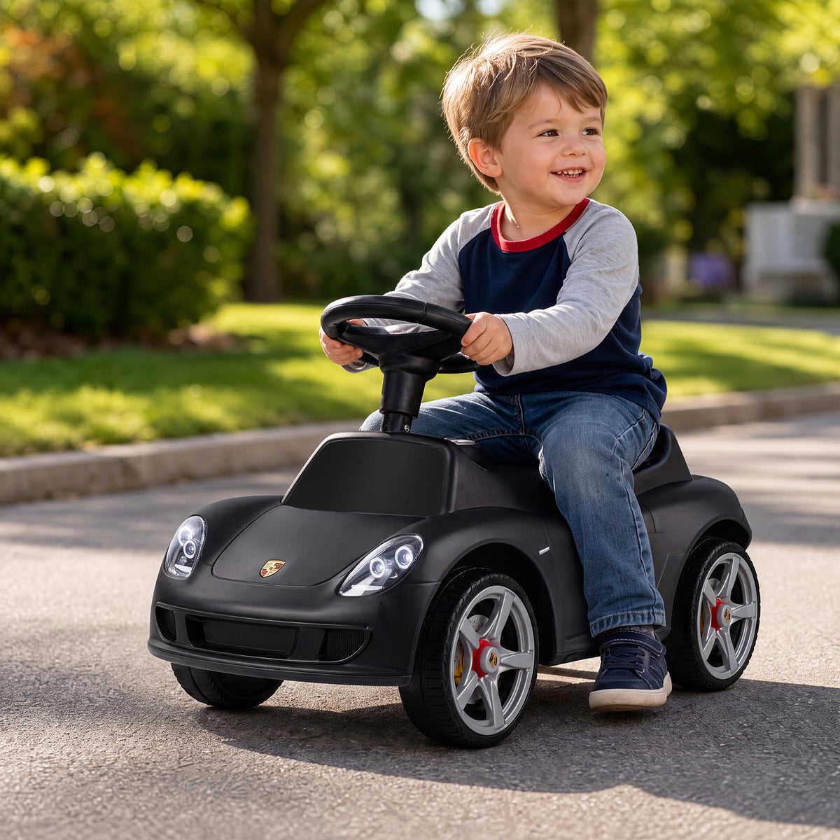 A happy toddler steers the black Porsche style sliding car on a paved path