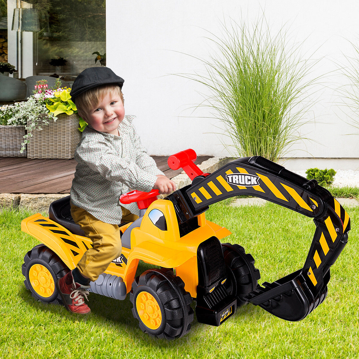 A young boy wearing a black cap rides the yellow and black digger toy on a green lawn with plants
