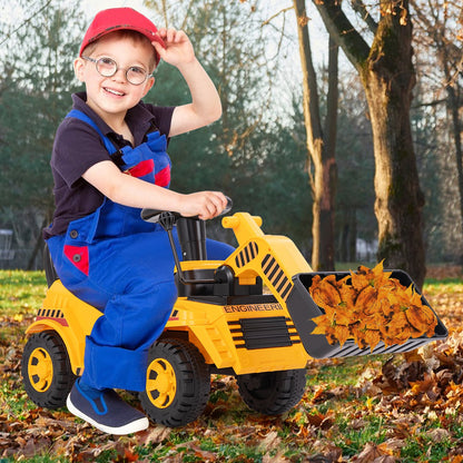 A child in a red cap and blue overalls plays with the yellow ride on digger, scooping leaves with the front bucket