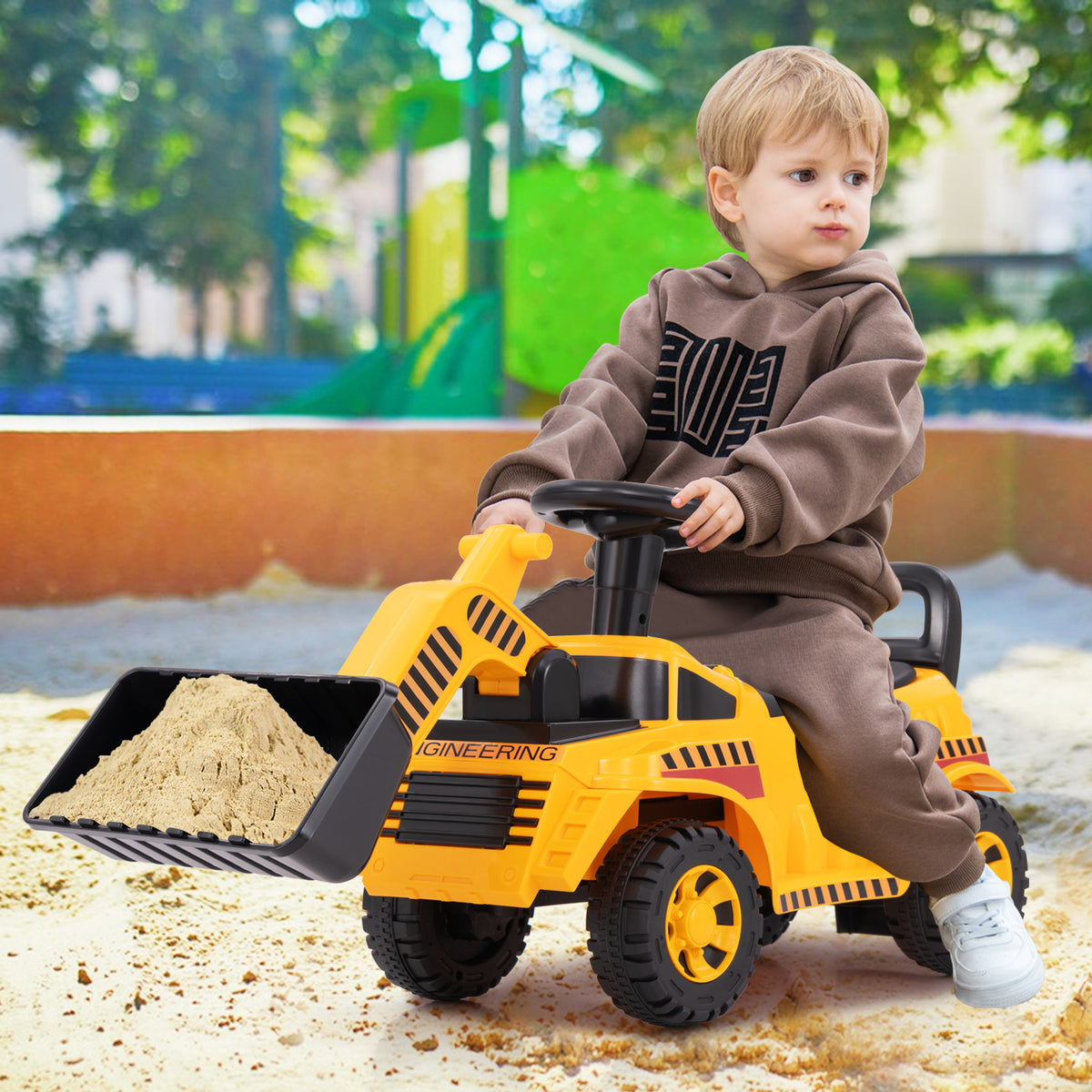 A young boy sits on the yellow ride on digger toy, pushing sand with the front bucket
