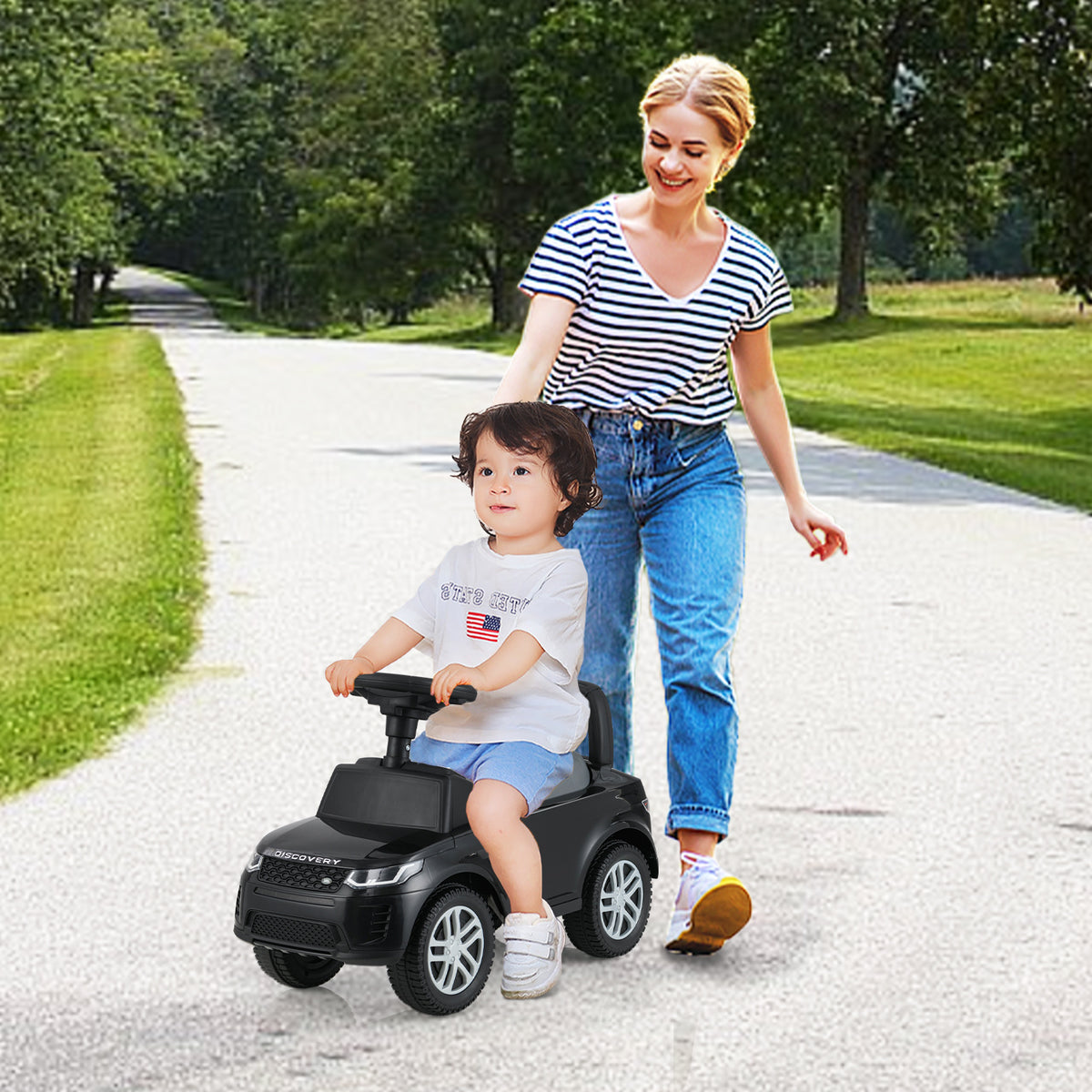 A woman pushes the black push and pedal ride on toy car with a toddler sitting on it outdoors