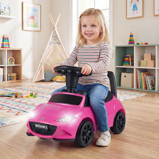 A happy toddler sits on the pink Mazda ride on toy in a playroom