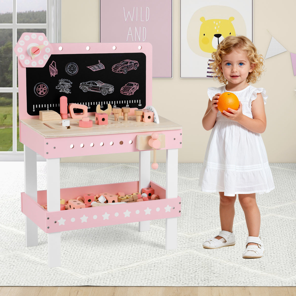 A young girl holding an orange stands next to the pink wooden play workbench