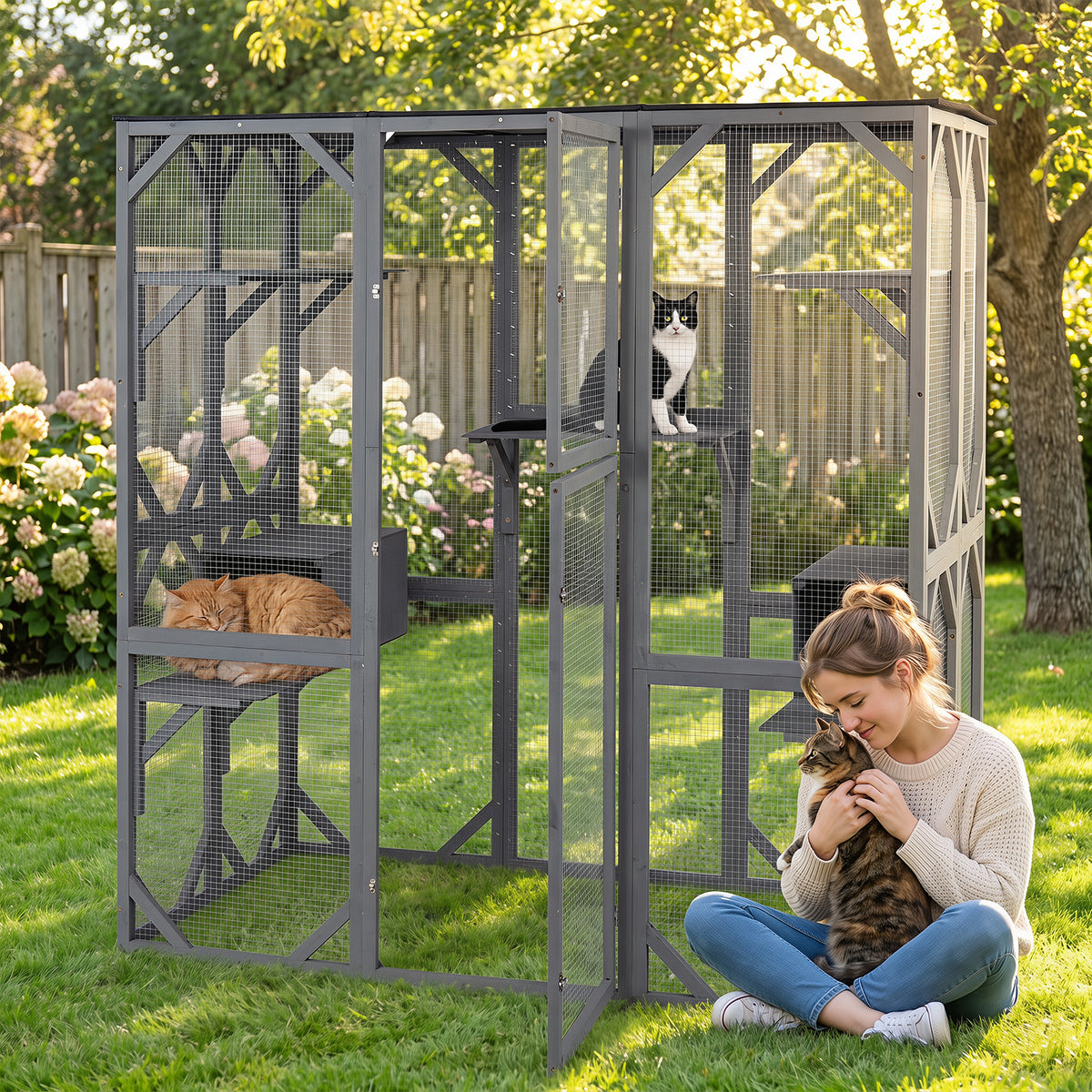 A woman sits in a garden holding a cat next to the outdoor cat enclosure