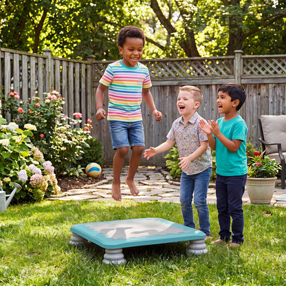 Three young boys laugh and jump on the blue square trampoline in a sunny garden