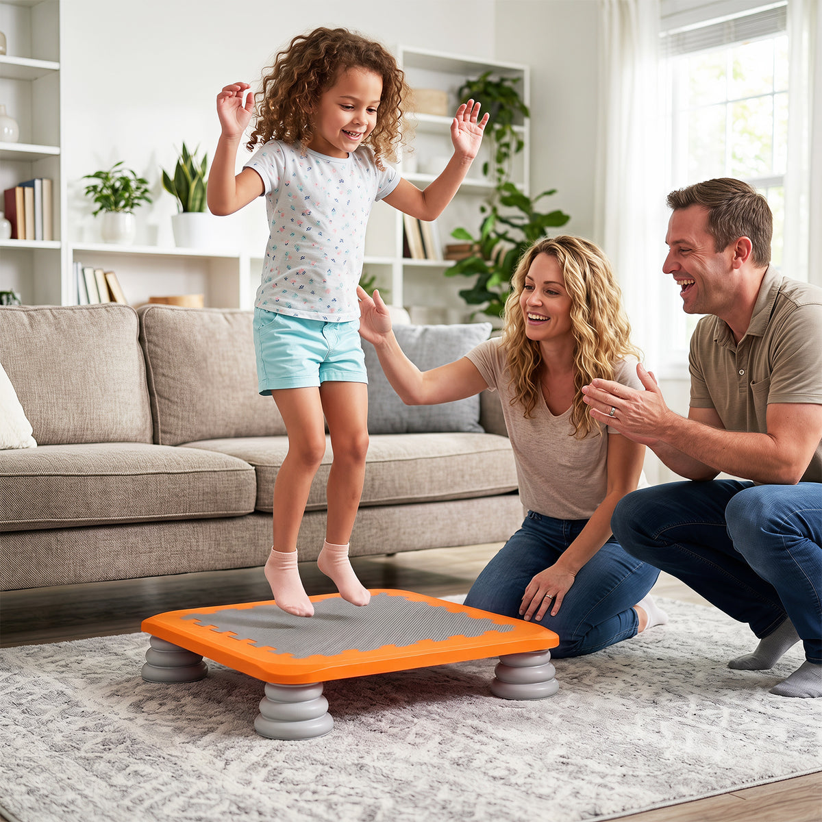 A young girl stands on an orange and gray balance board while her parents watch and cheer in a bright living room setting.