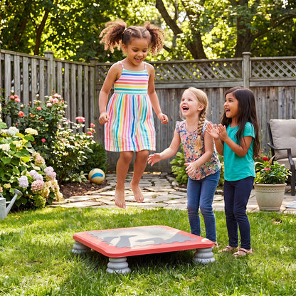Three young girls enjoy jumping on the red mini trampoline in a sunny garden