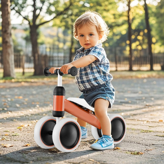 A toddler sits on the orange balance bike outdoors on a paved path