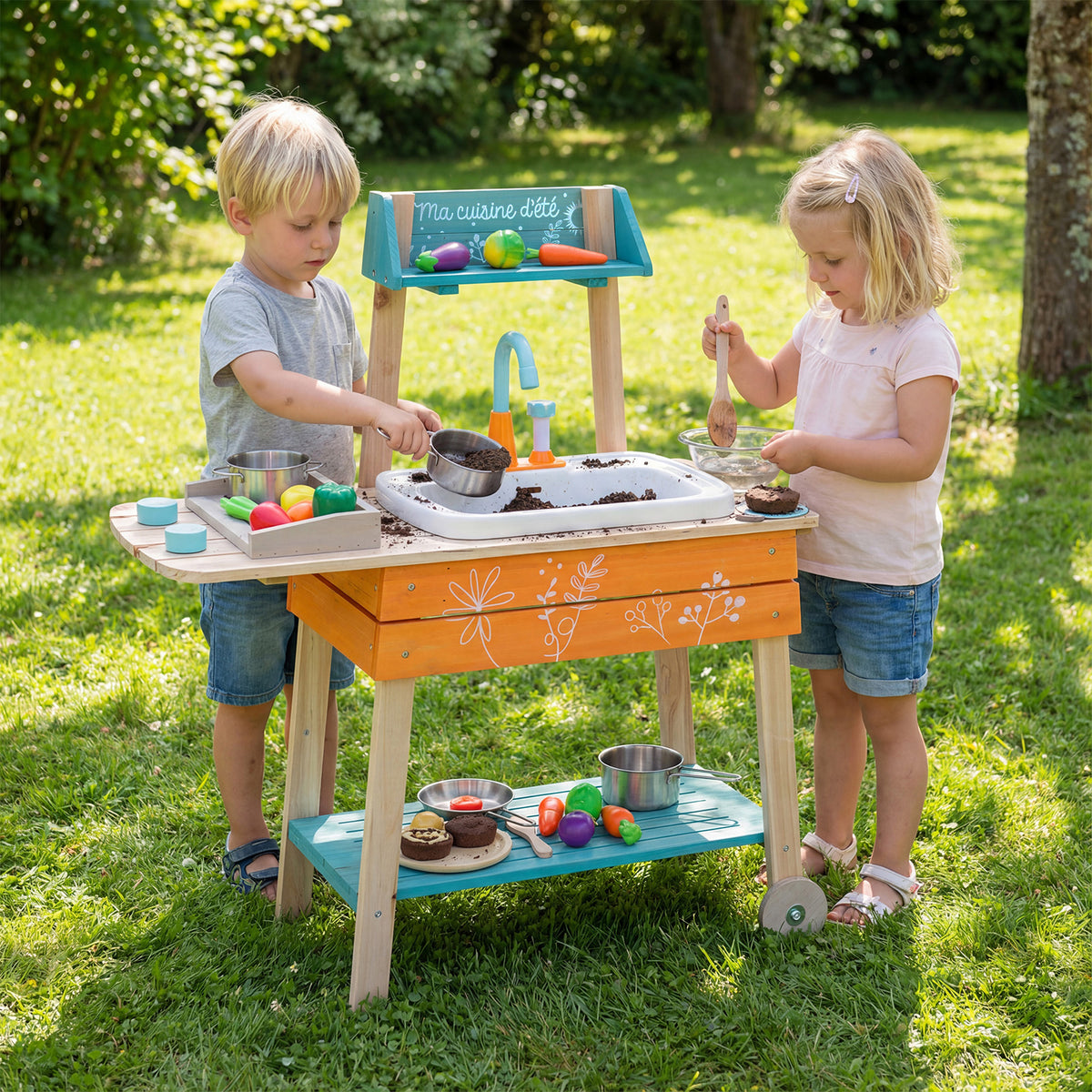 Two young children playing with the wooden play kitchen set in a sunny garden setting