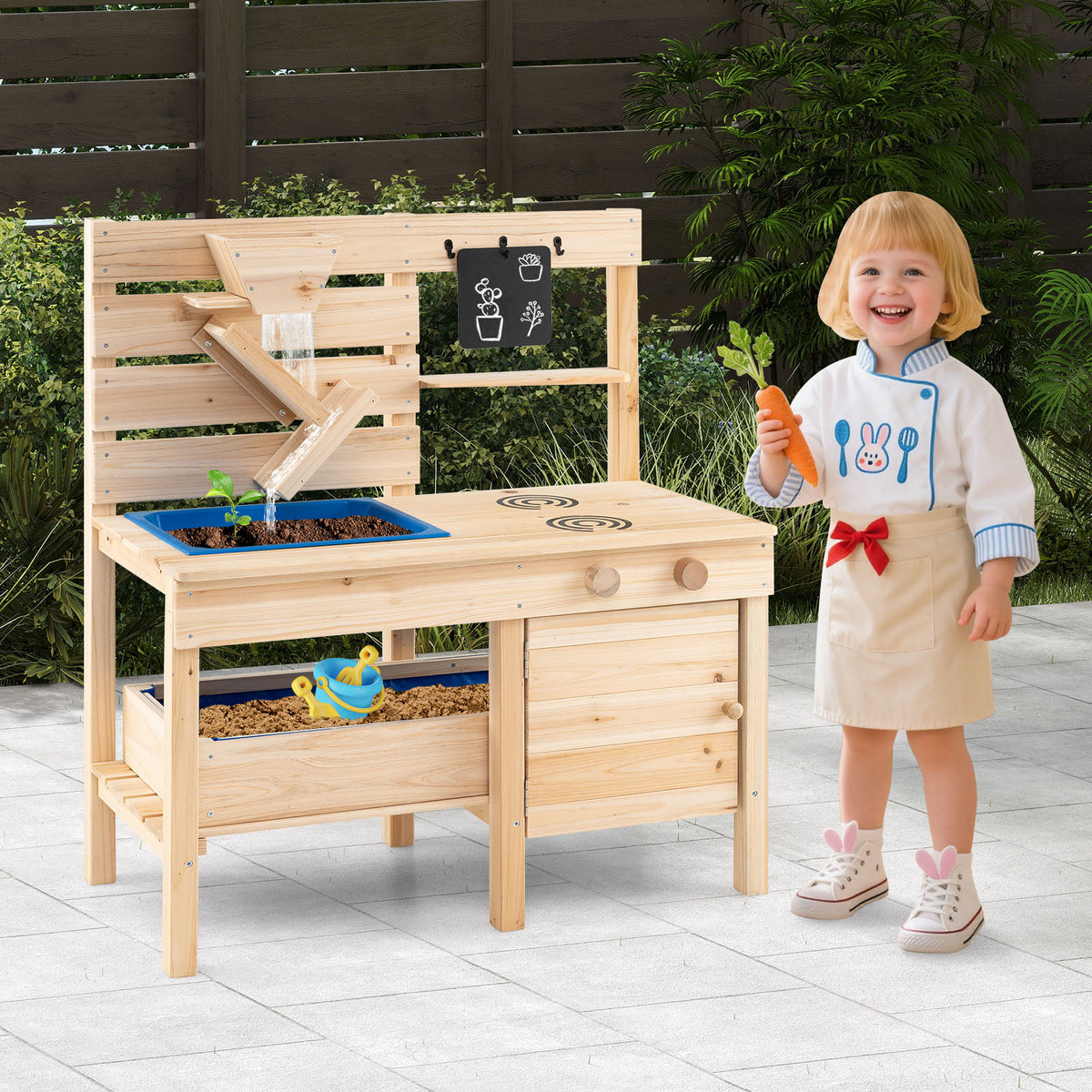 A young girl in a chef's outfit holds a carrot next to the play kitchen