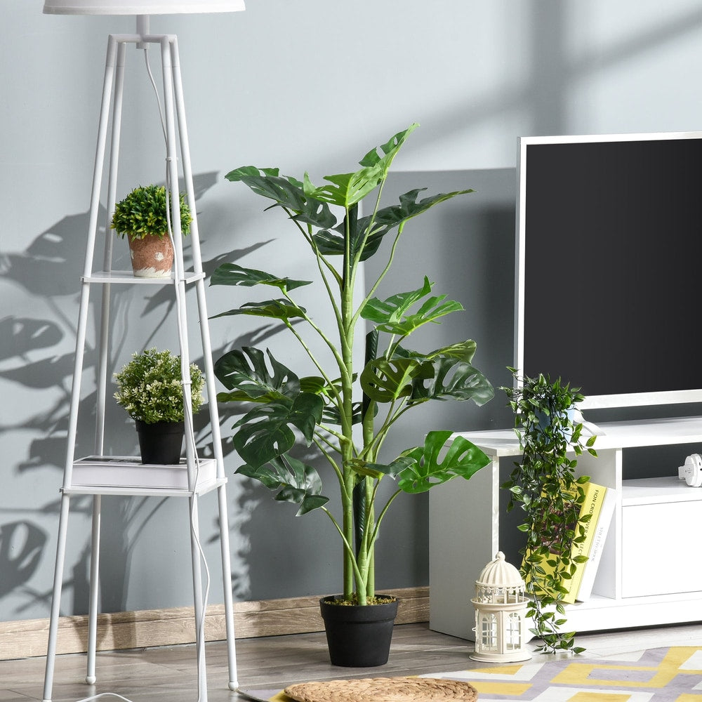 The artificial cheese plant is shown in a modern living room setting next to a television and a floor lamp