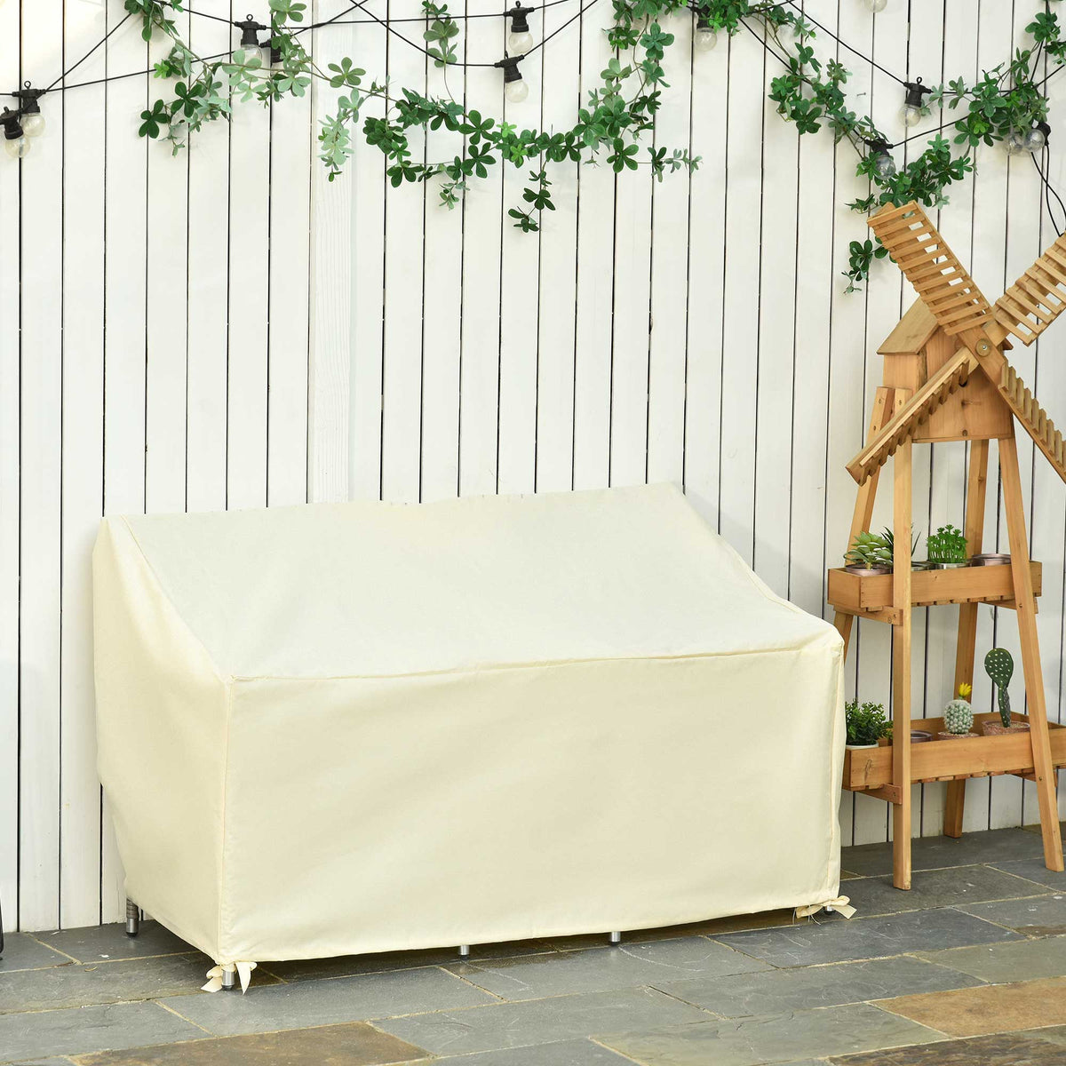 The beige outdoor sofa protector is shown on a patio with a wooden fence and plants