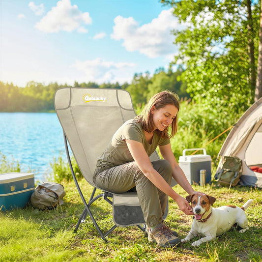 A woman and a dog relax on a folding camp chair next to a lake with a tent and cooler