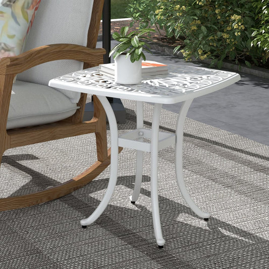 The white square garden table placed on a patio next to a rocking chair, with a plant on top