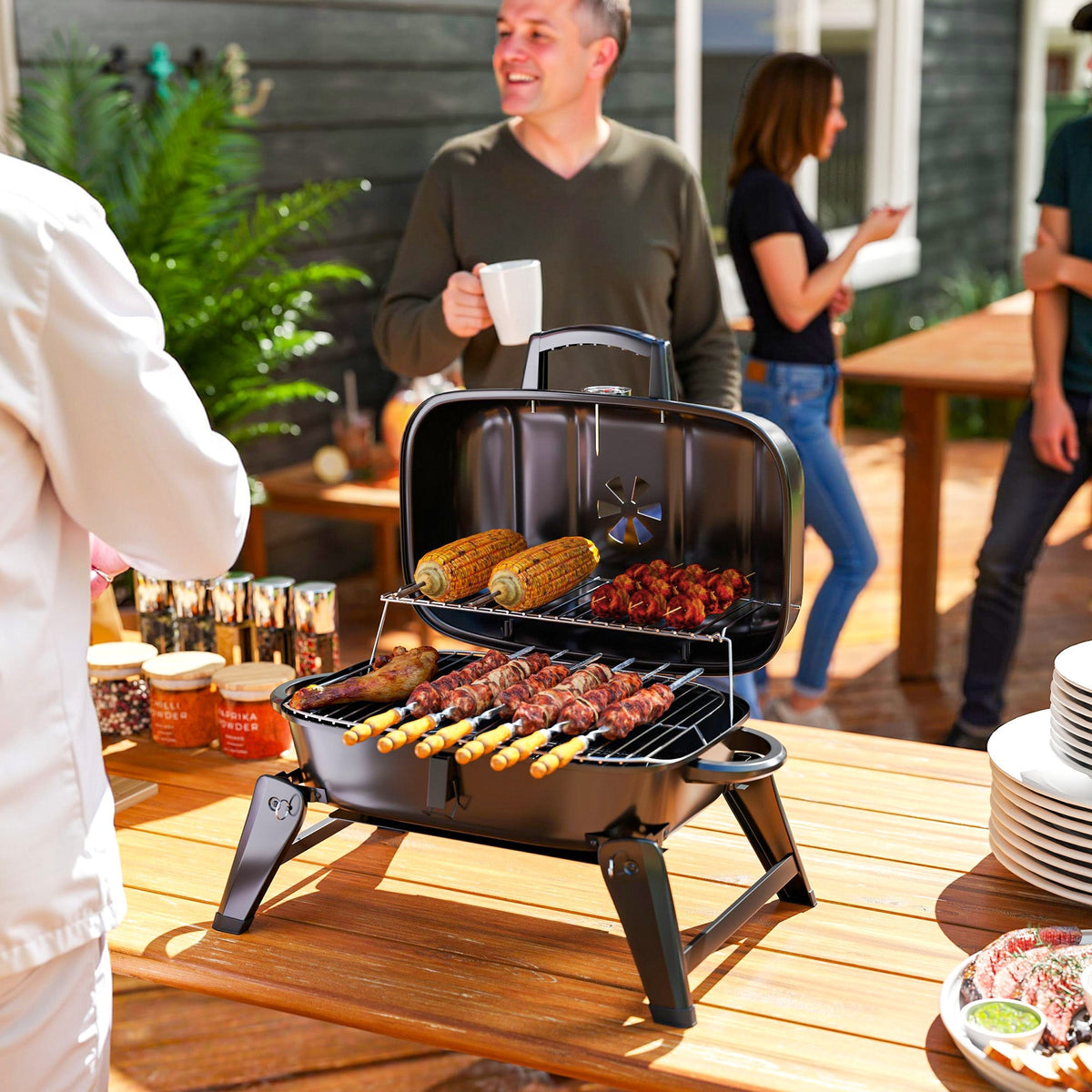The charcoal grill placed on a wooden table with food cooking while people socialise nearby