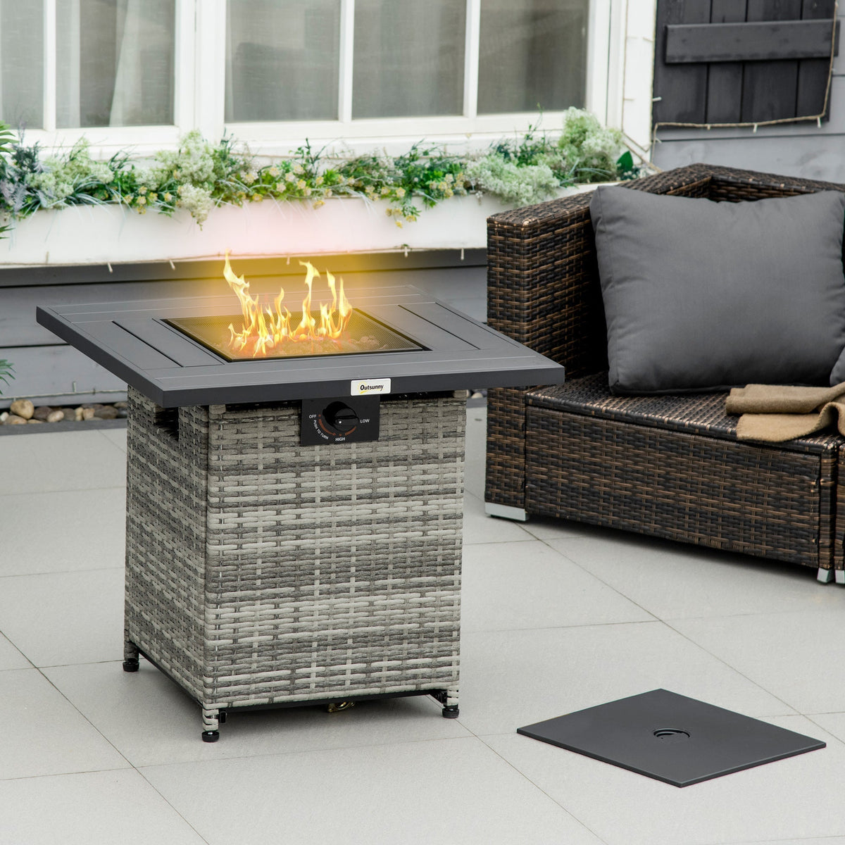The gas fire pit lit with flames on a patio next to a dark rattan sofa with a removed lid