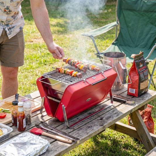 A red portable charcoal barbecue grill cooking skewers of meat and vegetables outdoors