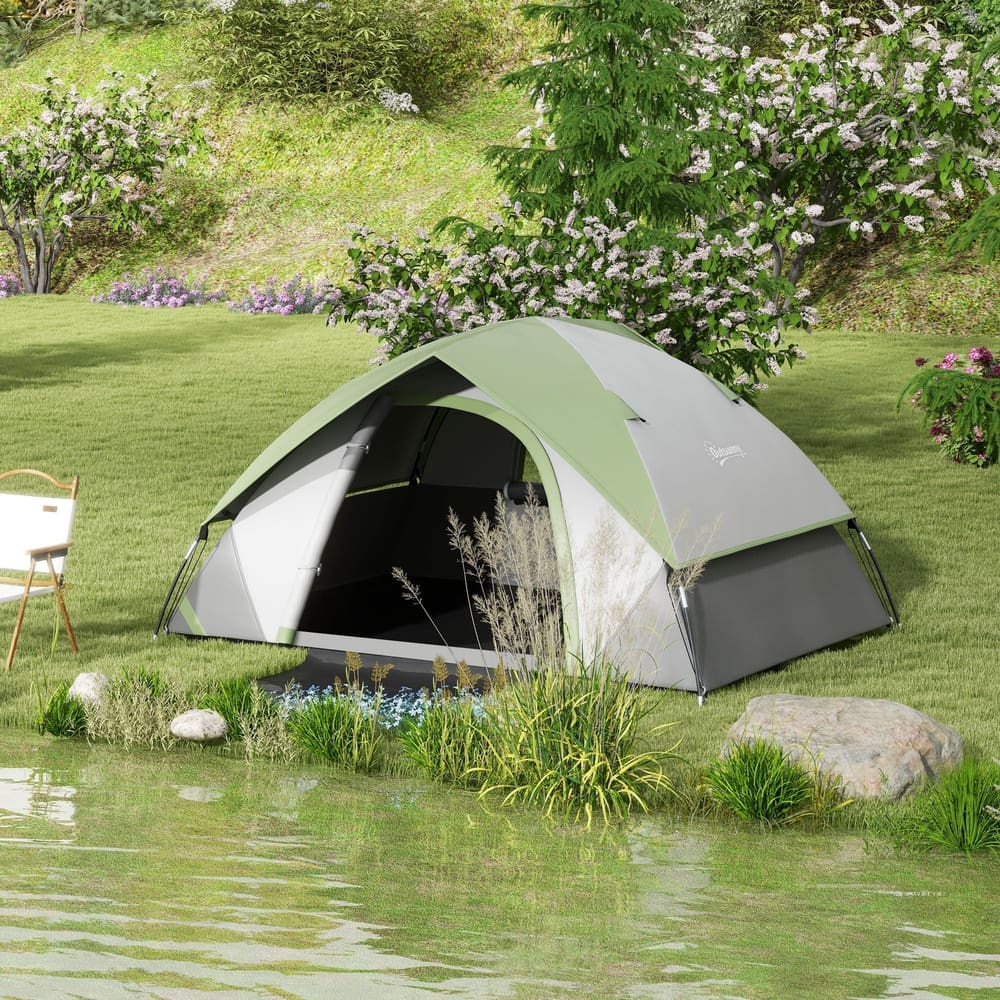 The green and grey camping tent pitched by a lake with trees and bushes in the background