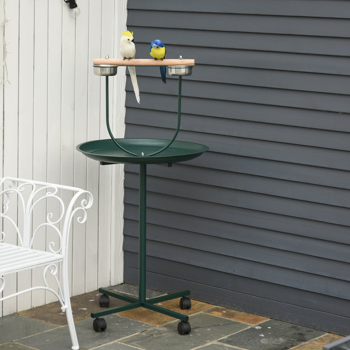 A green bird perch stand with four wheels, shown outdoors with two decorative birds on the perch