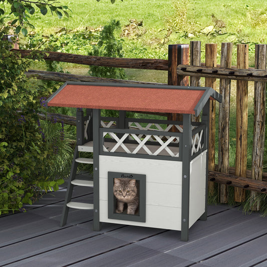 A grey tabby cat sits inside a white and grey outdoor cat house with a red roof and stairs