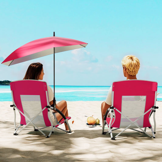 A pink beach chair with an umbrella attached, used by two people sitting on a sandy beach by the sea