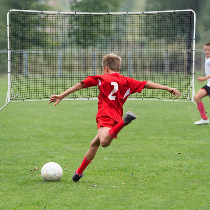 Young boy in a red football kit kicking a ball towards the rebounder net on a grass field