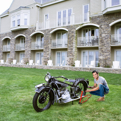 Man placing a vintage motorcycle onto a red rear wheel lift stand on grass