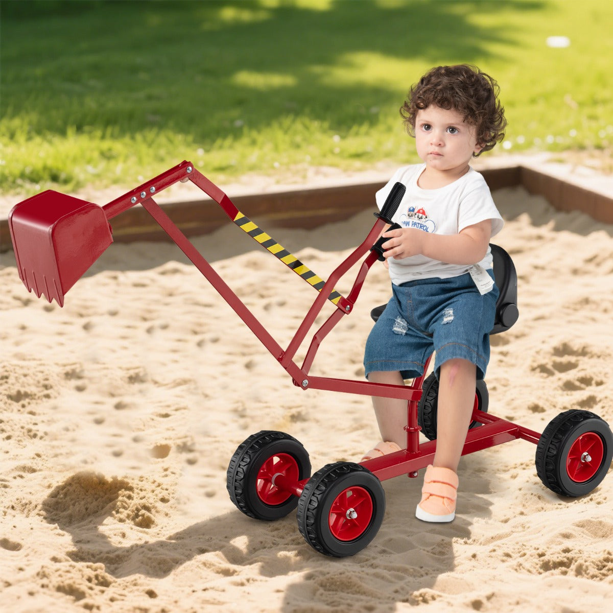 A young child sits on the red sand digger toy in a sandbox, operating the shovel