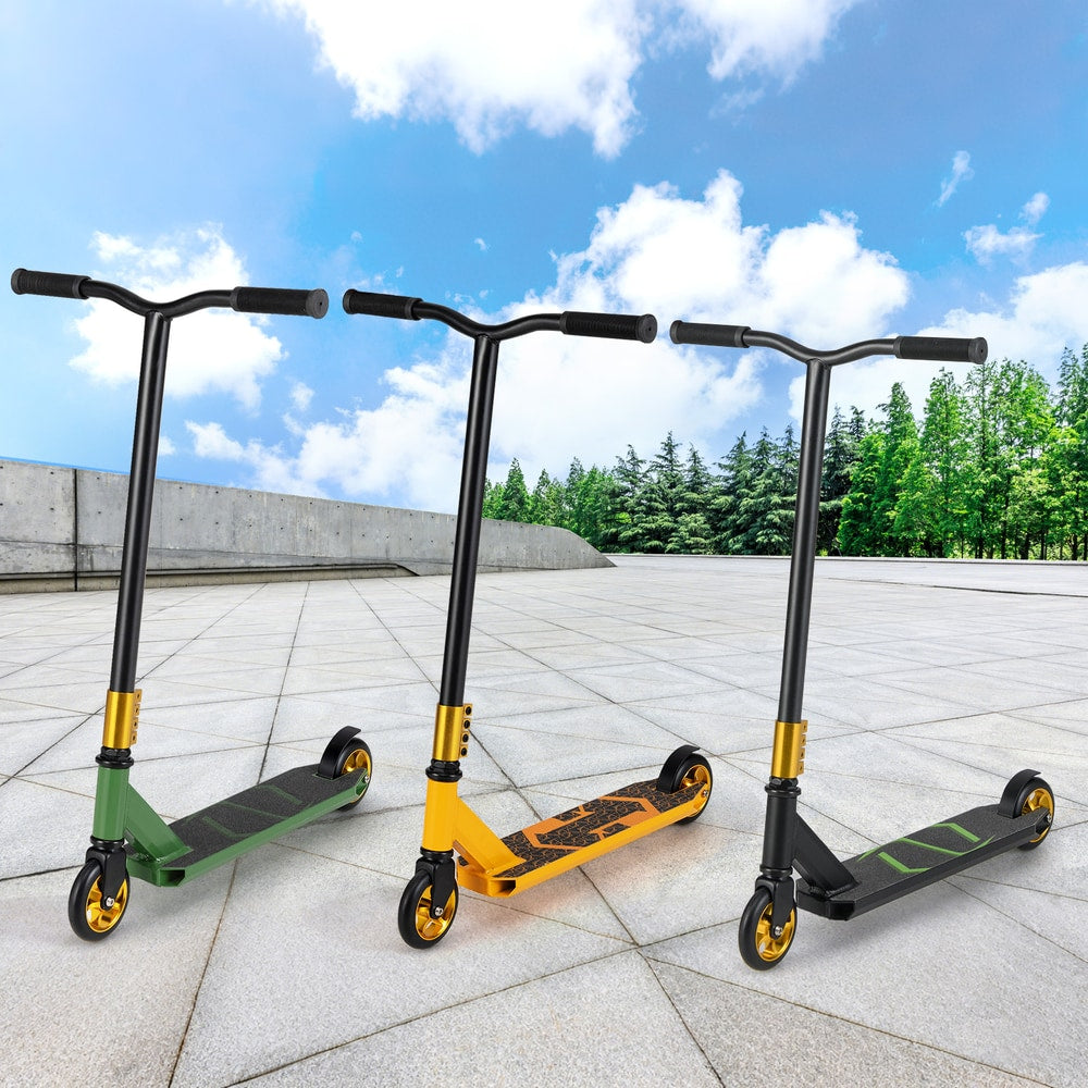 Three trick scooters in green, yellow, and black on a paved surface with trees and sky in the background