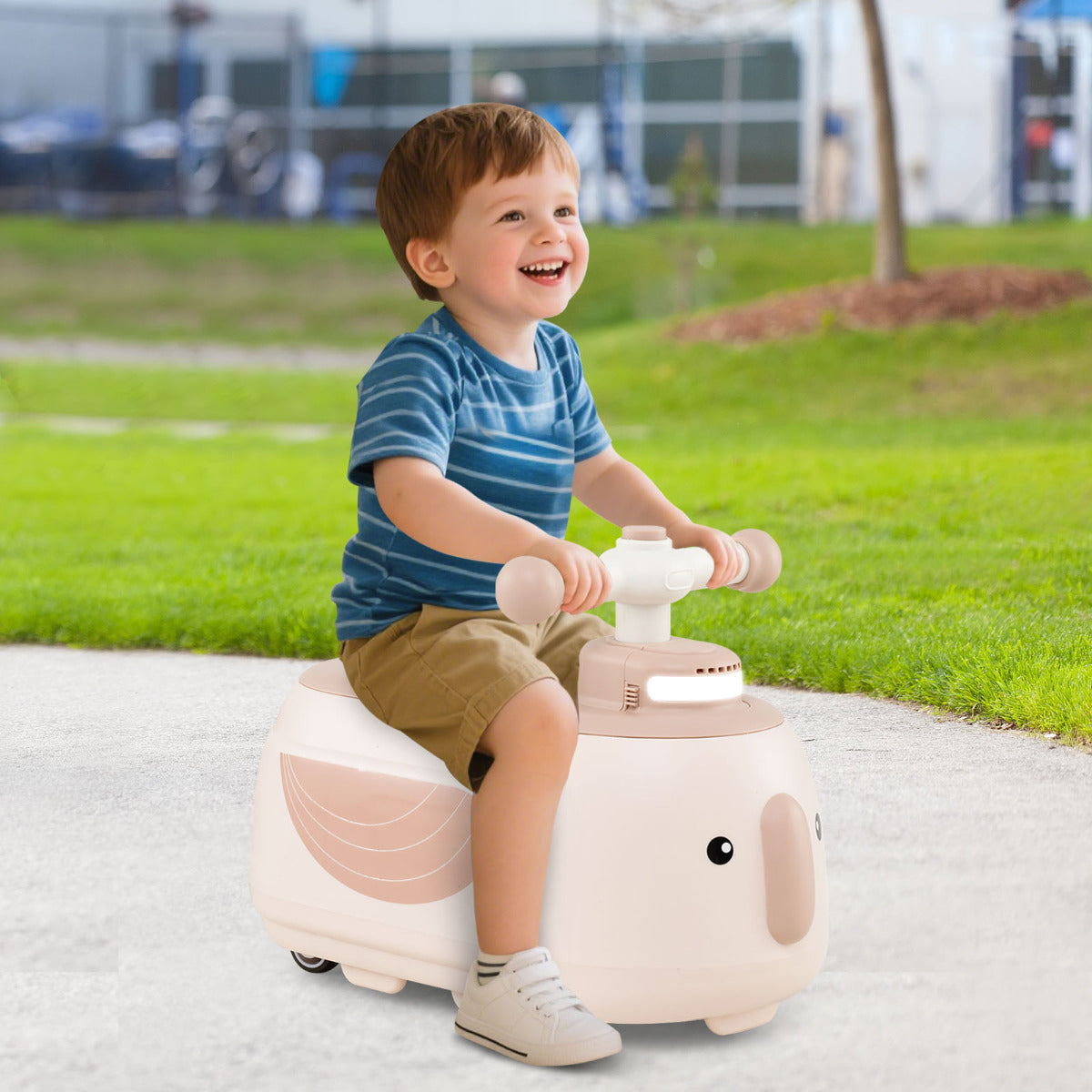 A smiling toddler boy rides the beige push and pedal toy with trailer outdoors on grass