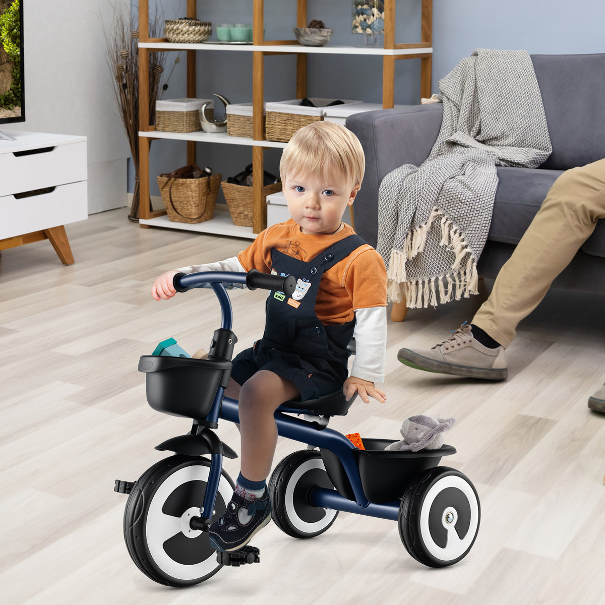 A young boy sits on the navy push and pedal ride on toy indoors with an adult's legs visible