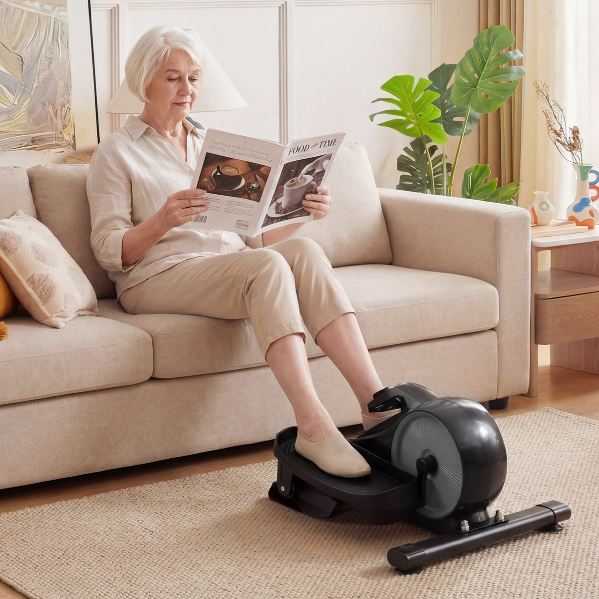 An older woman using the under desk elliptical pedal exerciser while sitting on a sofa reading