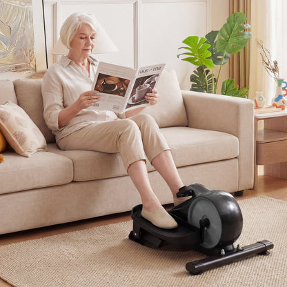 An older woman using the under desk elliptical pedal exerciser while sitting on a sofa reading